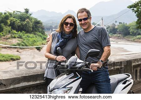 Stock Photograph - A couple stand posing by a motorcycle with a mountainous landscape in the distance; Luang Prabang, Luang Prabang Province, Laos. Fotosearch