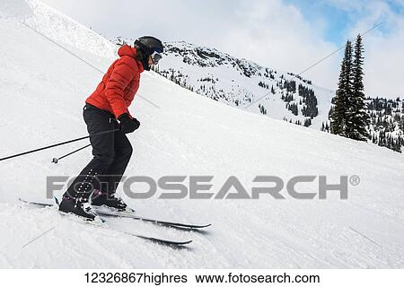 Stock Photograph - A downhill skier in a red coat skiing on the snow covered Rocky Mountains; Whistler, British Columbia, Canada. Fotosearch