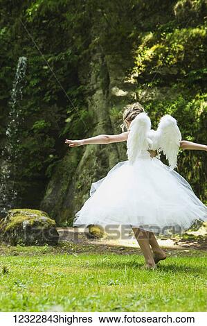 Stock Photograph - A girl with blond hair wearing an angel costume with feather wings walks on lush grass in a park; Oregon, United States of America. Fotosearch
