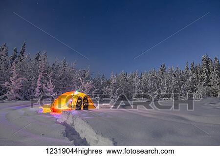 Stock Photograph - A glowing tent covered in string lights sits in the middle of an s0wy spruce forest, s0wshoes in the deep s0w outside the tent, moonlight casting shadows on a clear winter night, interior Alaska; Gakona, Alaska, United States of America. Fotosearch