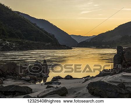 Stock Photograph - A golden sunset reflects into the tranquil water with silhouetted mountains; Luang Prabang Province, Laos. Fotosearch