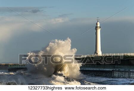 Stock Photograph - A large wave crashing against the wall on the shoreline below a lighthouse; Sunderland, Tyne and Wear, England. Fotosearch