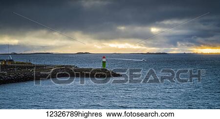 Stock Photograph - A lighthouse sits on the water's edge with a golden sun setting over the ocean under the clouds; Nordland, Norway. Fotosearch
