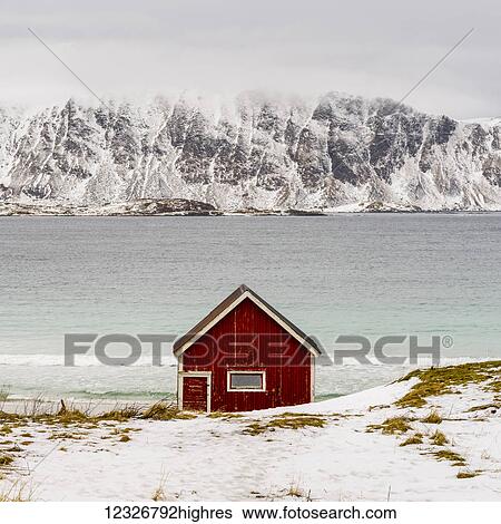 Stock Photograph - A lone red cabin at the water's edge with rugged, snow covered mountains across the water; Lofoten Islands, Nordland, Norway. Fotosearch