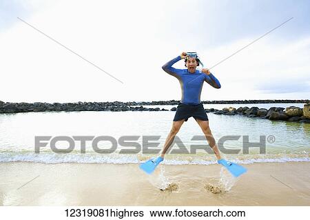 Stock Photograph - A man in s0rkel gear stands at the water's edge on Lydgate Beach and jumps in the air; Kauai, Hawaii, United States of America. Fotosearch