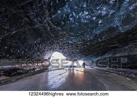 Stock Photograph - A man stands near the entrance to a tunnel beneath the ice of Canwell Glacier with the colors of sunset reflecting off the ice in mid-winter; Alaska, United States of America. Fotosearch