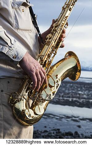 Stock Photograph - A man stands with a saxophone on the beach at the water's edge; Alaska, United States of America. Fotosearch