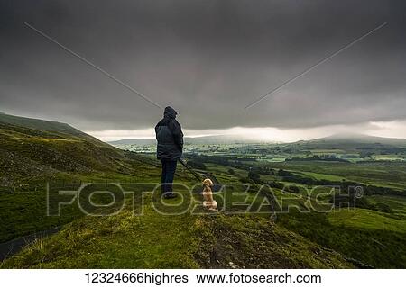 Stock Photograph - A man stands with his dog on a grassy hill looking out over the lush, green landscape under a stormy sky; North Yorkshire, England. Fotosearch