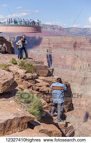 Stock Photograph - A native american worker photographs tourists at the Skywalk Viewpoint over the West Grand Canyon natural landscape area in Arizona, a popular tourist attraction operated by the indigenous native American people; Arizona, United States of America. Fotosearch