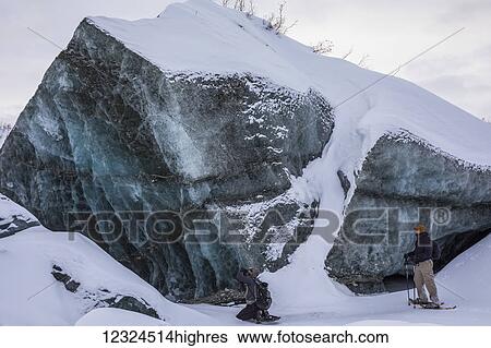 Stock Photograph - A pair of snowshoers observe a large chunk of ice near the terminus of Canwell Glacier in the Alaska Range; Alaska, United States of America. Fotosearch