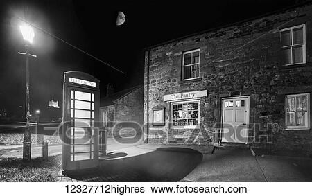 Stock Photograph - A phone booth illuminated by a street lamp at nighttime outside a shop; Bamburgh, Northumberland, England. Fotosearch