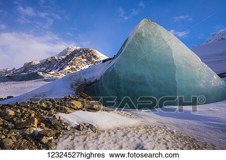 Stock Photograph - A piece of glacier ice juts out of the rocky, snow-covered moraine of Black Rapids Glacier in the winter; Alaska, United States of America. Fotosearch