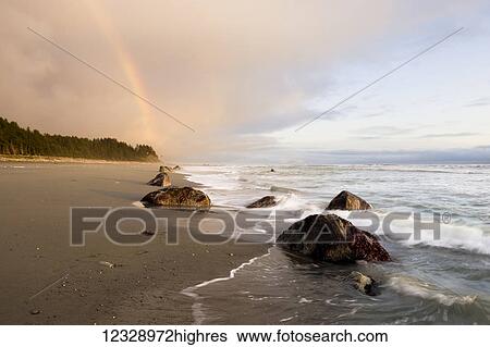 Stock Photograph - A rainbow glows in storm clouds over a beach and coastline; Alaska, United States of America. Fotosearch