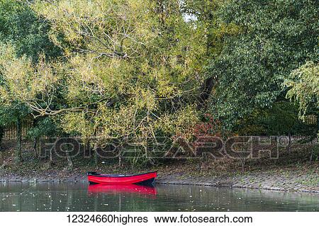 Stock Photograph - A red boat sits along the shoreline of tranquil water of River Tyne; Gateshead, Tyne and Wear, England. Fotosearch