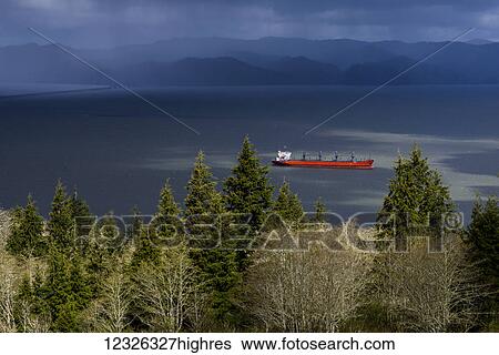 Stock Photograph - A red ship passes through Columbia river under dramatic storm clouds and silhouetted mountains in the background; Astoria, Oregon, United States of America. Fotosearch