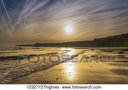 Stock Photograph - A sinking sun glows in the sky and reflects off the wet sand along the coast; South Shields, Tyne and Wear, England. Fotosearch