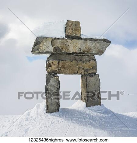 Stock Photograph - A stone inuksuk in the snow under a cloudy sky with a snow covered mountain in the background; Whistler, British Columbia, Canada. Fotosearch