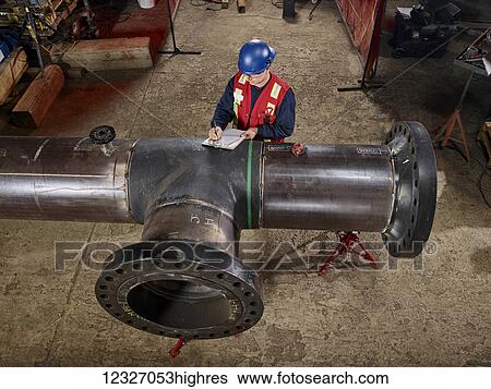 Stock Photograph - A tradesman stands at a large pipe and writes on a document; Edmonton, Alberta, Canada. Fotosearch