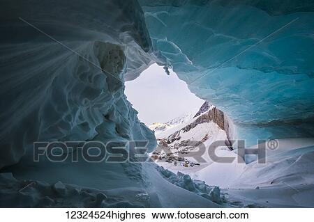 Stock Photograph - A wave-like formation of snow curls around the entrance of a small ice cave in the moraine of Black Rapids Glacier in winter; Alaska, United States of America. Fotosearch