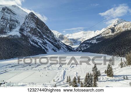 Stock Photograph - A wide angle view of Lake Louise from the upper floor of Chateau Lake Louise Resort in the winter surrounded by snow capped mountains and filled with skaters on the ice rinks carved into the frozen lake; Lake Louise, Alberta, Canada. Fotosearch