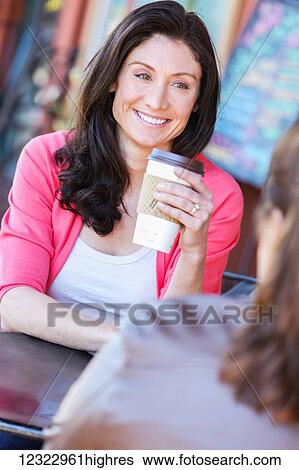 Stock Photograph - A woman sits at a coffee shop holding a paper cup of coffee while sitting across from a friend; Oregon, United States of America. Fotosearch