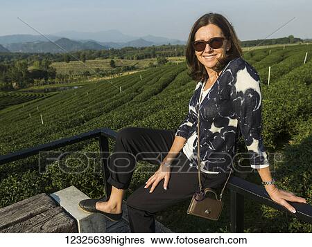 A woman sits on a railing with the slopes of a tea plantation in the background; Tambon Si Kham, Chang Wat Chiang Rai, Thailand View Large Photo Image Stock Photograph - A woman sits on a railing with the slopes of a tea plantation in the background; Tambon Si Kham, Chang Wat Chiang Rai, Thailand. Fotosearch