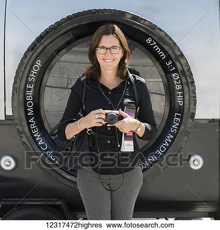 Stock Photograph - A woman stands holding her camera with a large image of a camera lens as the backdrop; Seoul, South Korea. Fotosearch