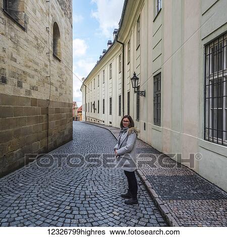 Stock Photograph - A woman stands posing on a narrow street with patterned paving stones in between buildings; Prague, Czech Republic. Fotosearch