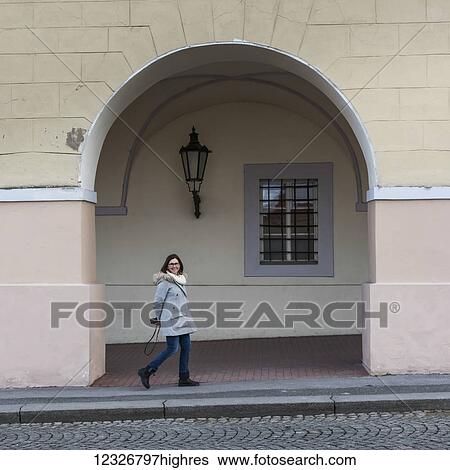 Stock Photograph - A woman strolls by a building under an archway with a smile, holding a camera behind her back; Prague, Czech Republic. Fotosearch