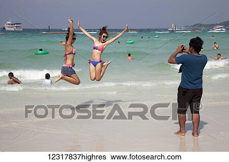 Stock Photograph - A woman takes a picture of two teenage girls leaping mid-air on the beach while swimming at the ocean; Pattaya, Thailand. Fotosearch