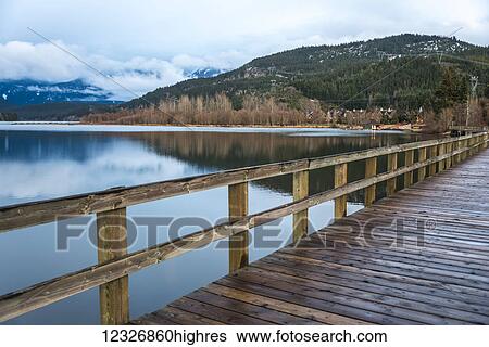 Stock Photograph - A wooden boardwalk along a tranquil lake with mountains along the shoreline; Whistler, British Columbia, Canada. Fotosearch