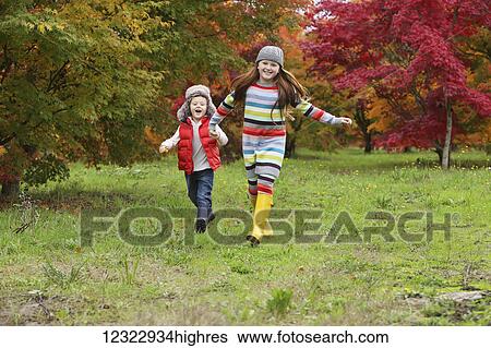 Stock Photograph - A young boy and girl wearing rubber boots and colourful clothing run across a field holding hands with trees in bright autumn colours in the background; Oregon, United States of America. Fotosearch