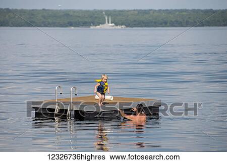 Stock Photograph - A young Caucasian girl prepares to jump off a floating dock into her dad's arms in Seneca Lake in upstate New York, the naval barge, no longer in service, is in the background; Dresden, New York, United States of America. Fotosearch
