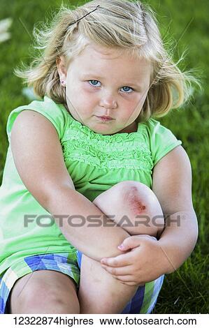 Stock Photograph - A young girl sits on the grass looking unhappy while holding her knee that has a scrape on it; Oregon, United States of America. Fotosearch