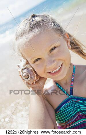 A young girl smiling, holding and listening to the shell sound by her ear at the beach; Honolulu, Oahu, Hawaii, United States of America View Large Photo Image Stock Photograph - A young girl smiling, holding and listening to the shell sound by her ear at the beach; Honolulu, Oahu, Hawaii, United States of America. Fotosearch