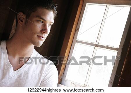 Stock Photograph - A young man stands looking out a window; Oregon, United States of America. Fotosearch