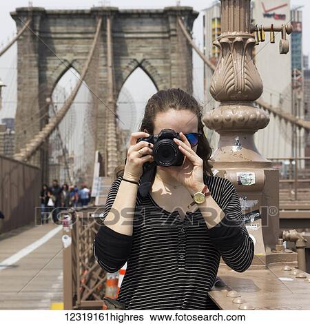 Stock Photograph - A young woman holds a camera to take a photograph, pointing at the camera, on the Brooklyn Bridge; New York City, New York, United States of America. Fotosearch
