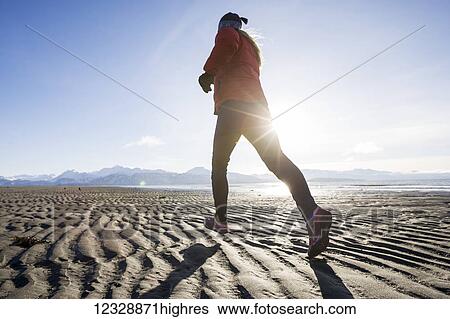 Stock Photograph - A young woman jogs on the wet beach; Homer, Alaska, United States of America. Fotosearch