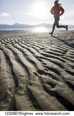 Stock Photograph - A young woman jogs on the wet beach; Homer, Alaska, United States of America. Fotosearch