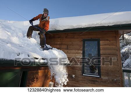 Stock Photograph - A young woman removes snow from the roof of her house; Homer, Alaska, United States of America. Fotosearch