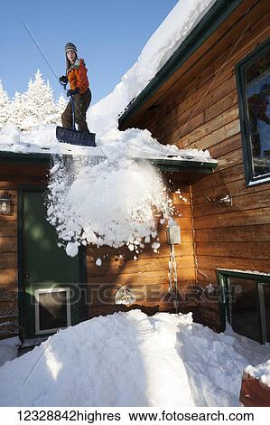 Stock Photograph - A young woman removes snow from the roof of her house; Homer, Alaska, United States of America. Fotosearch