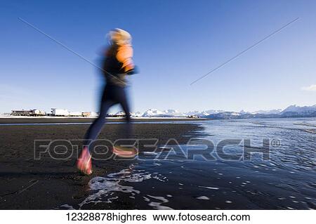 Stock Photograph - A young woman running along the water's edge; Homer, Alaska, United States of America. Fotosearch