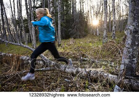 Stock Photograph - A young woman running over logs on a forest floor in a forest; Homer, Alaska, United States of America. Fotosearch