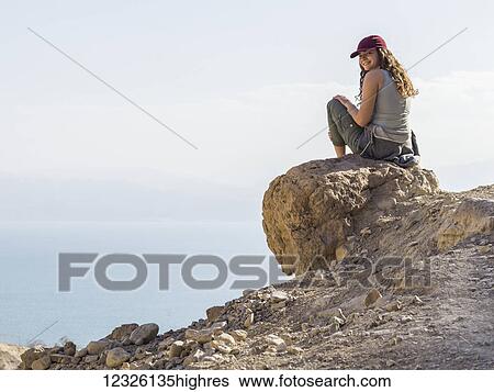A young woman sits on a rock looking back and posing for the camera with a cloud covered valley down below, Ein Gedi Nature Reserve, Dead Sea District; South Region, Israel View Large Photo Image Stock Photograph - A young woman sits on a rock looking back and posing for the camera with a cloud covered valley down below, Ein Gedi Nature Reserve, Dead Sea District; South Region, Israel. Fotosearch
