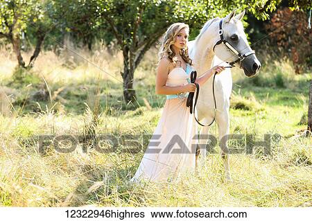 Stock Photograph - A young woman standing in a flowing dress with a white horse; Oregon, United States of America. Fotosearch