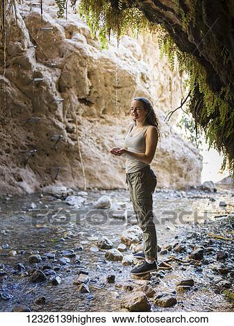 Stock Photograph - A young woman stands on a rock along a stream at Dodim Cave, Ein Gedi Nature Reserve; South District, Israel. Fotosearch