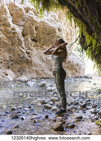 Stock Photograph - A young woman stands on a rock and looks up along a stream at Dodim Cave, Ein Gedi Nature Reserve; South District, Israel. Fotosearch