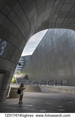 Stock Photograph - A young woman stands under a curved wall using her smart phone, Dongdaemun Design Plaza; Seoul, South Korea. Fotosearch