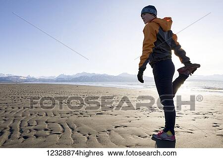 Stock Photograph - A young woman stretches on the wet beach before running; Homer, Alaska, United States of America. Fotosearch