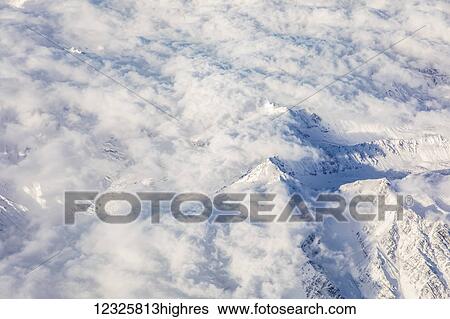 Stock Photograph - Aerial view of a snowy mountain peak rising above low altitude clouds in the Denali Mountain range, the sun casting shadows in the low valleys below, South-central Alaska; Alaska, United States of America. Fotosearch